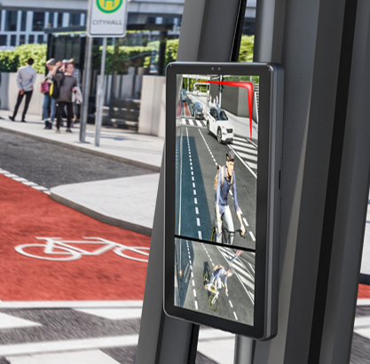 The view of a monitor mounted on the A column of a commercial vehicle. The monitor shows the scene right next to the vehicle, usually the blind spot. The image shows a cyclist from two different angles. To the front, there is a red bike line and a bus stop.